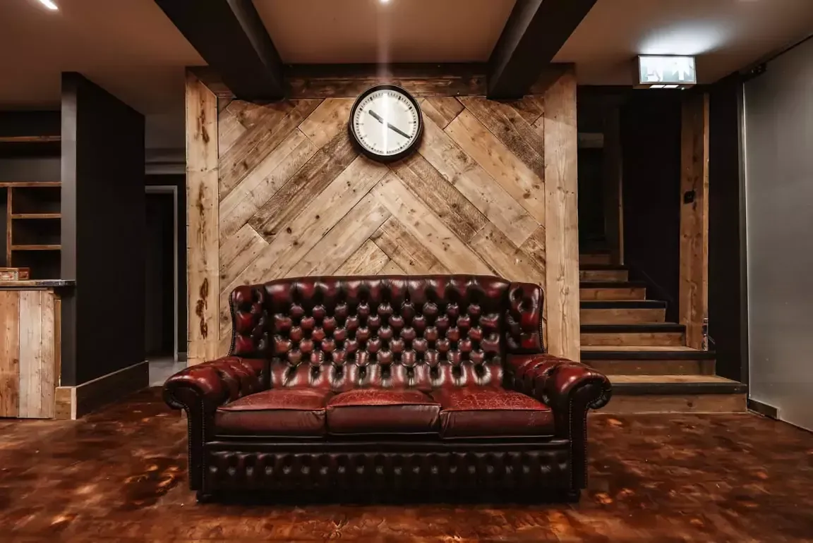 A red leather couch is sitting in front of a wooden wall with a clock on it.