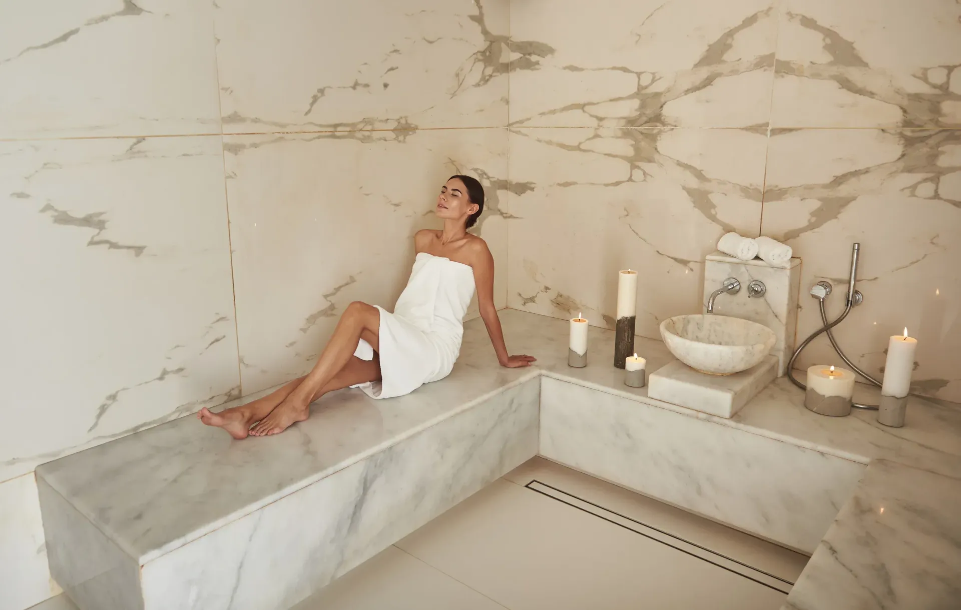 Una mujer envuelta en una toalla blanca se relaja en un spa con paredes de mármol, velas y un lavabo.