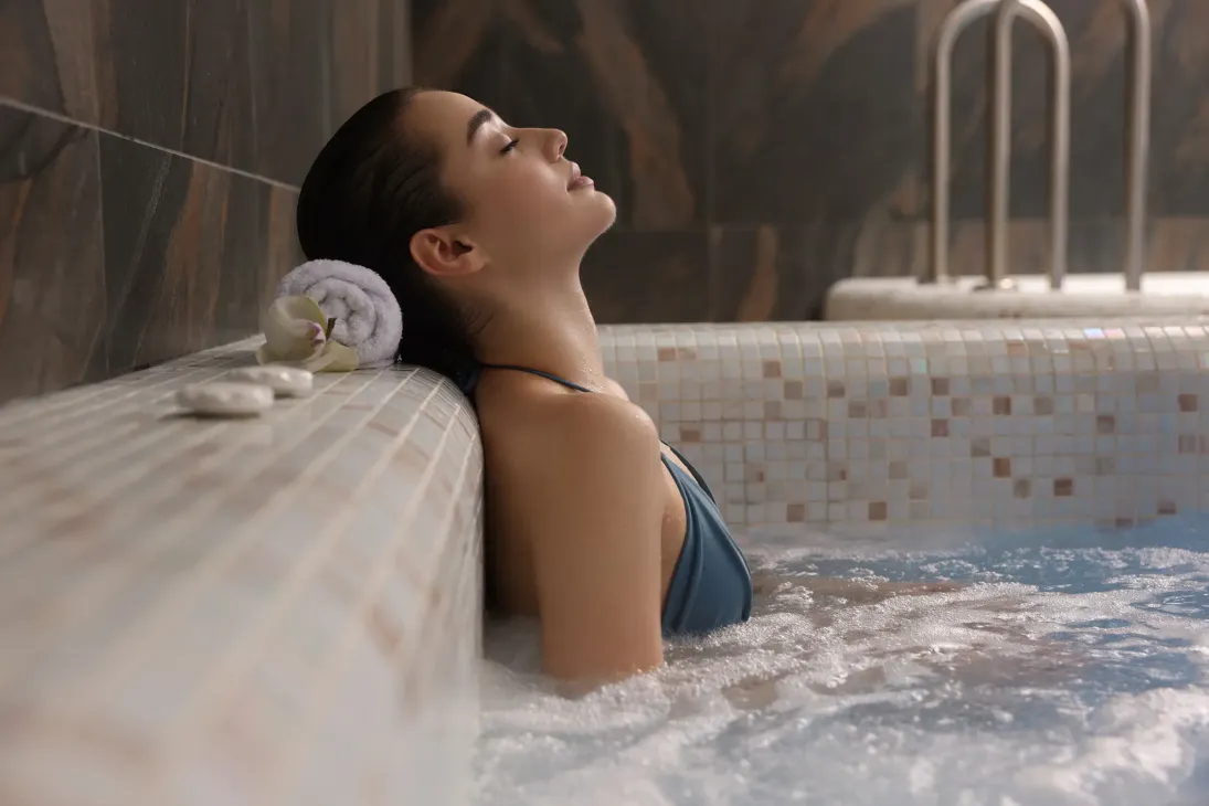 Mujer relajándose en un jacuzzi, reclinada con los ojos cerrados, toalla blanca y piedras cerca.