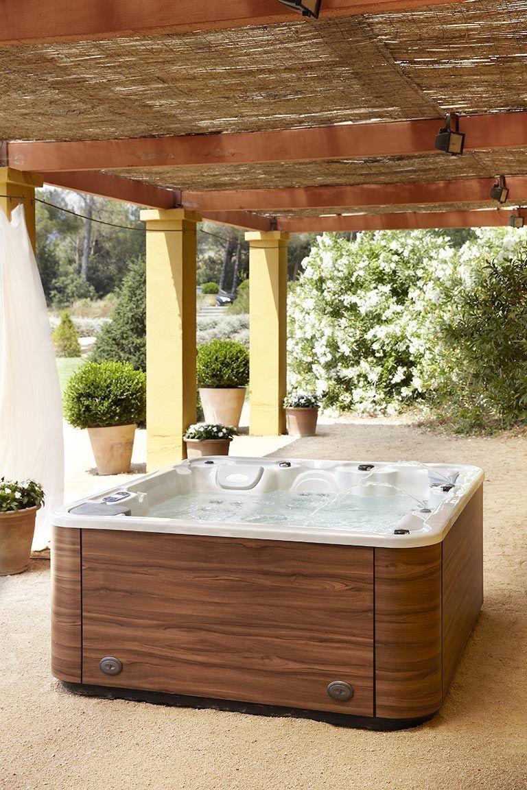 Jacuzzi al aire libre bajo una pérgola con revestimiento de madera, suelo de color canela y exuberantes plantas verdes.