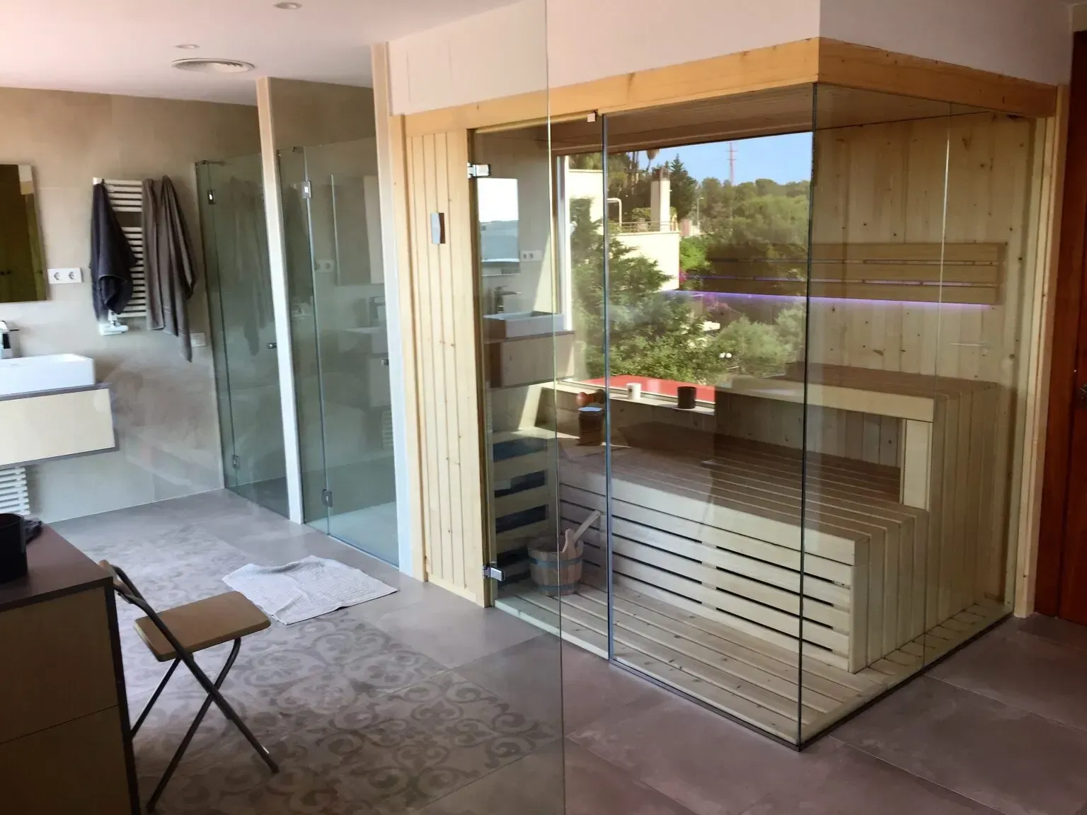 Baño moderno con sauna acristalada. Bancos de madera, paredes de cristal y vistas a los árboles del exterior.