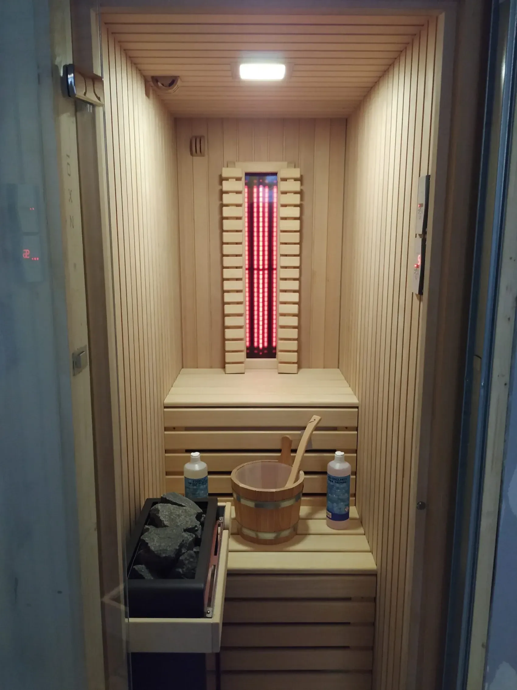 Interior de una sauna de madera con banco, luz infrarroja, cubo de agua y calentador de piedras.
