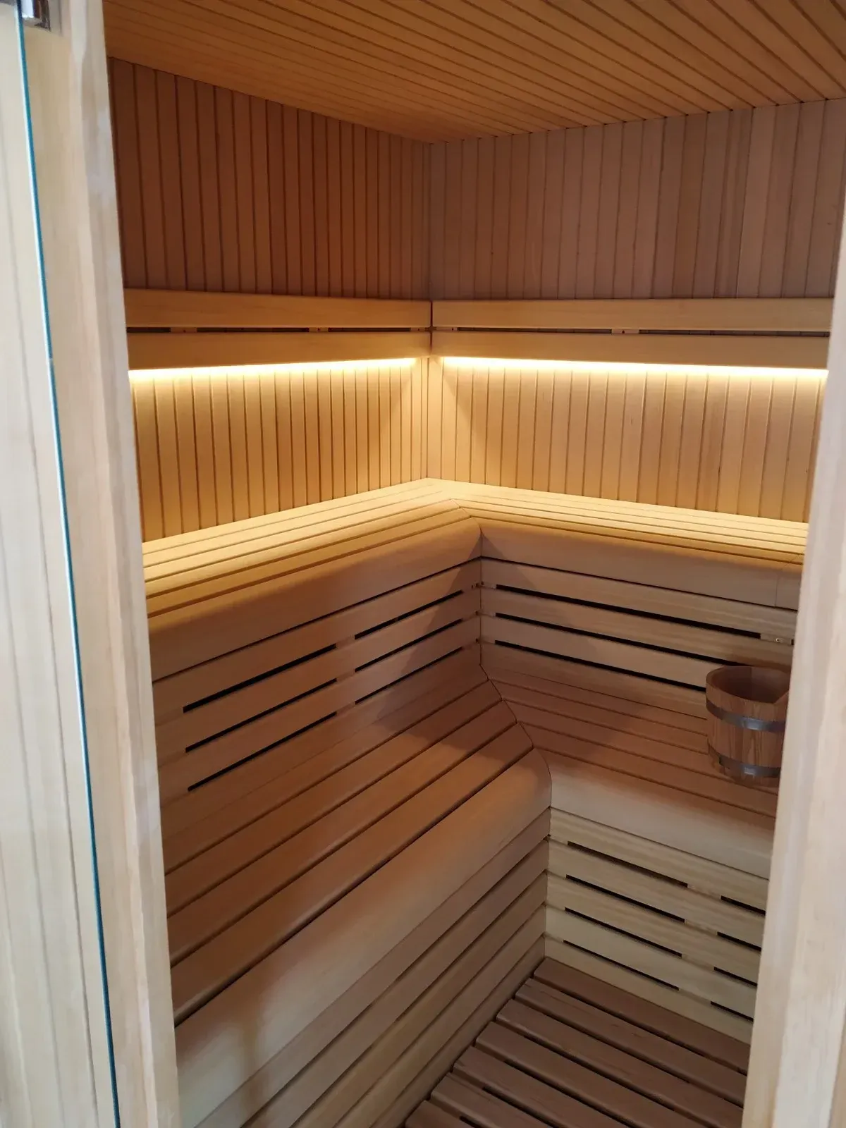 Interior de sauna de madera con bancos, iluminación LED y un cubo de madera.