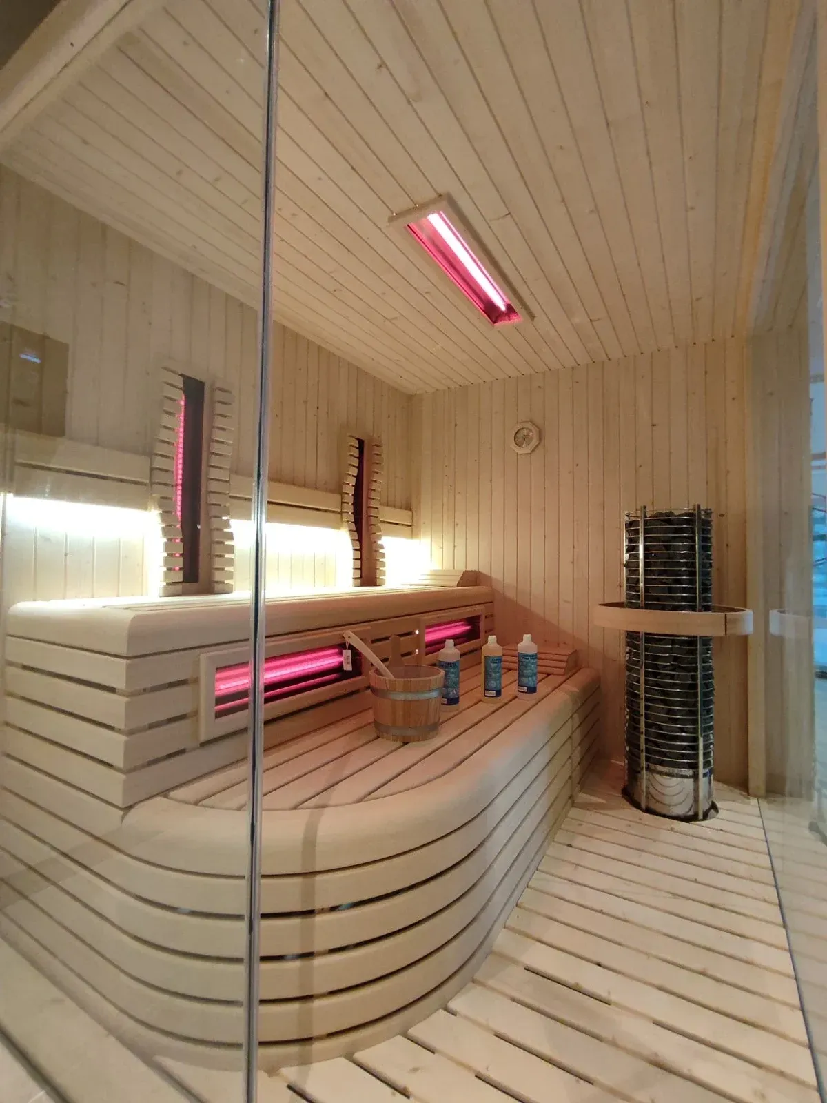 Interior de sauna de madera con lámparas de calor rojas, bancos y un calentador cilíndrico.