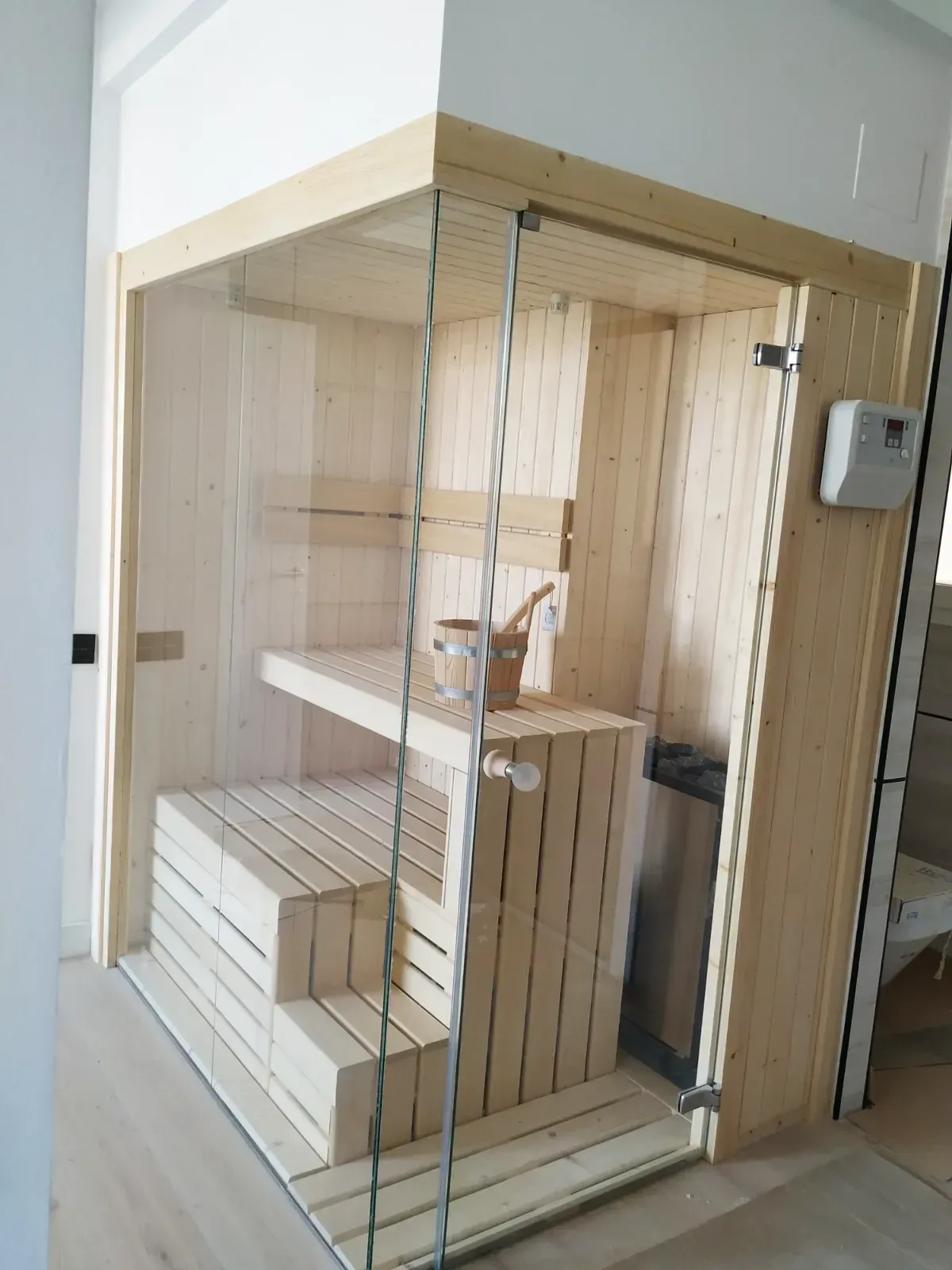 Una sauna de madera clara con puerta de cristal, bancos y balde.