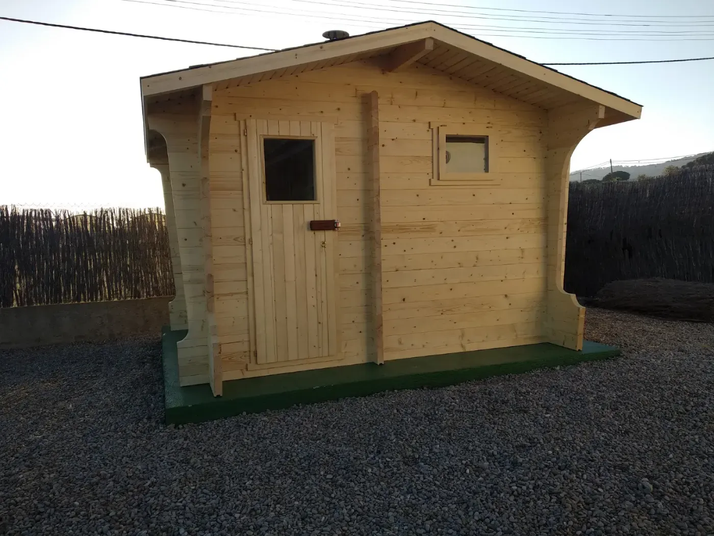Edificio de sauna de madera con puerta y ventana, situado sobre grava, con una base verde y una valla oscura al fondo.