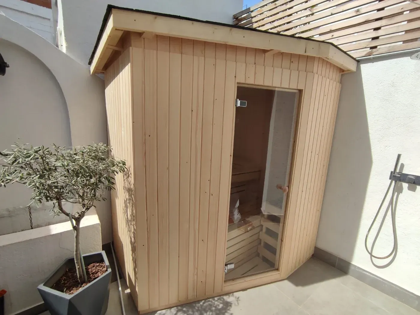 Sauna de madera en un patio, junto a un pequeño árbol en una maceta, con ducha exterior.