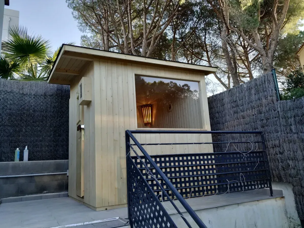 Edificio de sauna de madera con ventana, construido sobre una losa de hormigón con barandilla y escalones