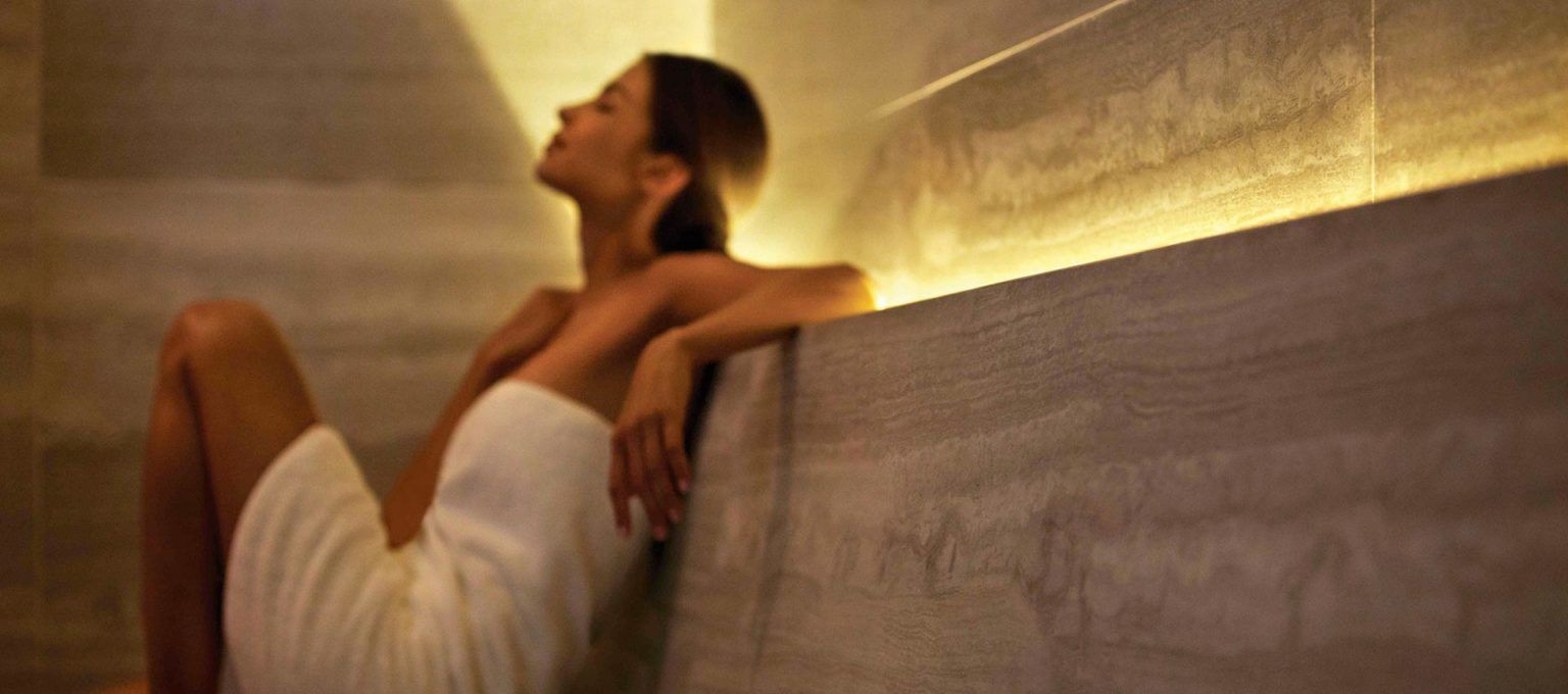 Mujer con una toalla blanca relajándose en un baño de vapor, apoyada contra un muro de piedra. Iluminación cálida.