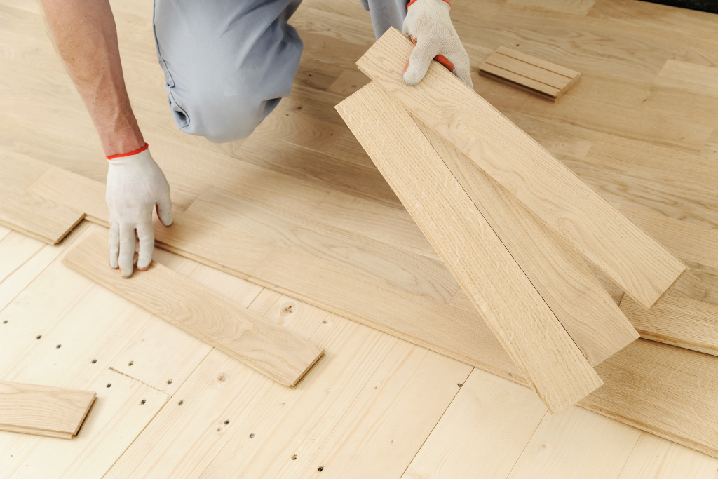 Personne à genoux, installant des planches de parquet; mains dans des gants blancs, bois blond.