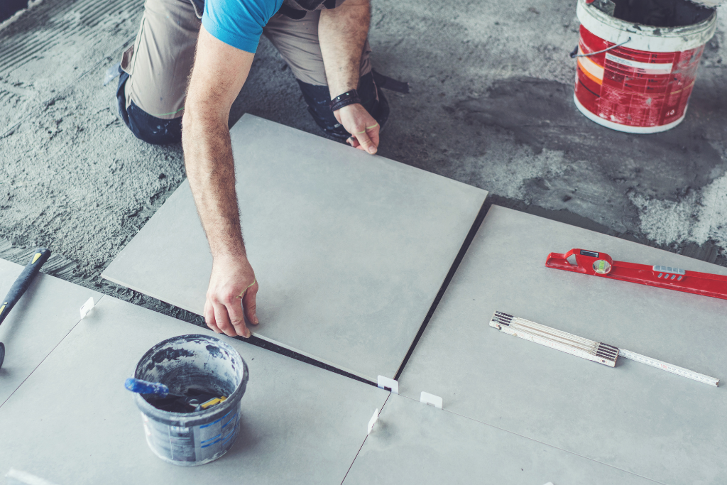 Personne posant des carreaux gris sur un sol en béton, à l'aide d'entretoises et d'outils.
