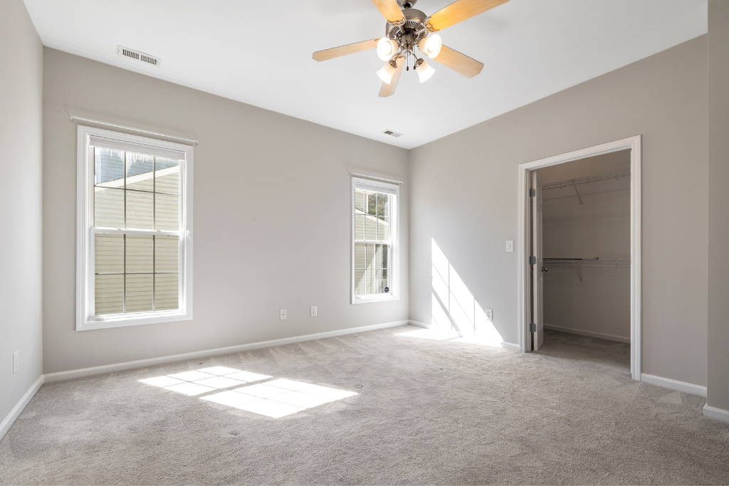 Chambre vide avec deux fenêtres, porte de placard et ventilateur de plafond, avec des couleurs neutres et de la moquette.