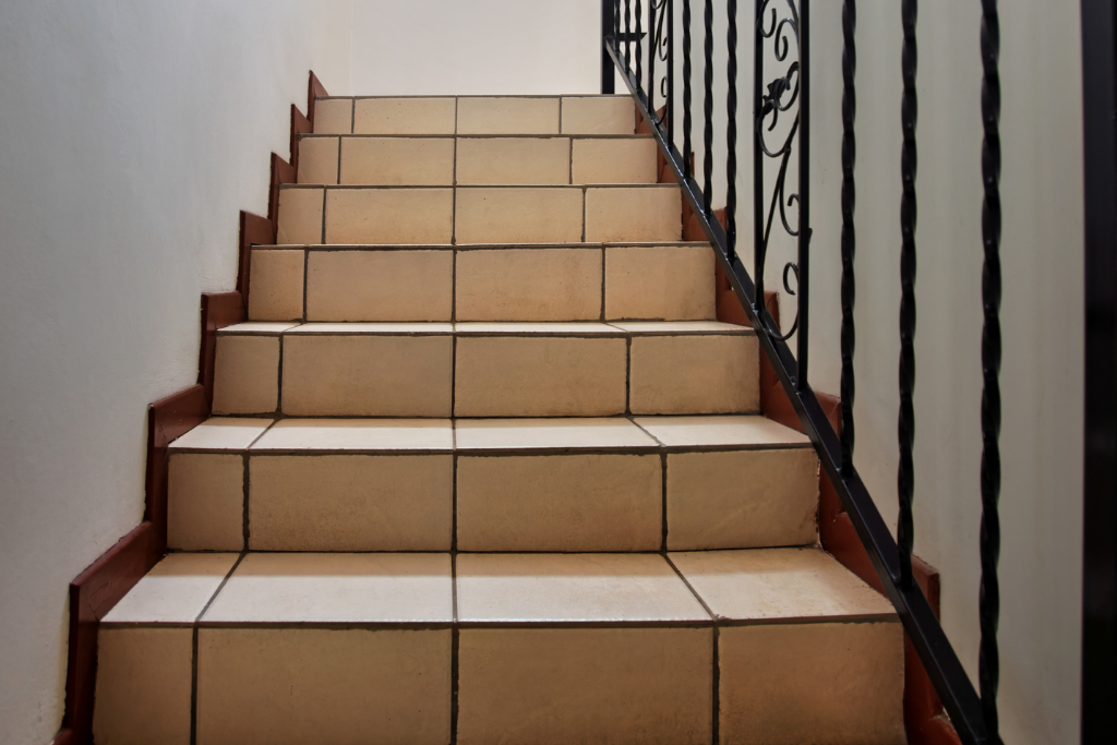 Escalier avec carrelage beige et rampe décorative noire, montant contre un mur blanc.