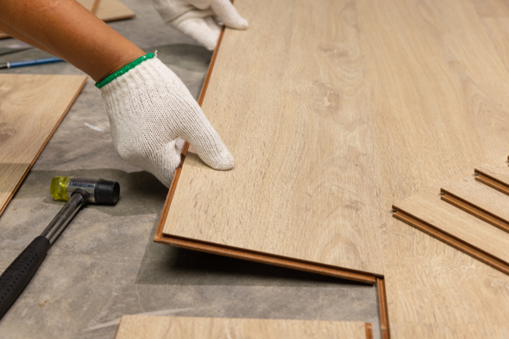 Personne portant des gants installant un parquet en bois brun clair ; marteau à proximité.