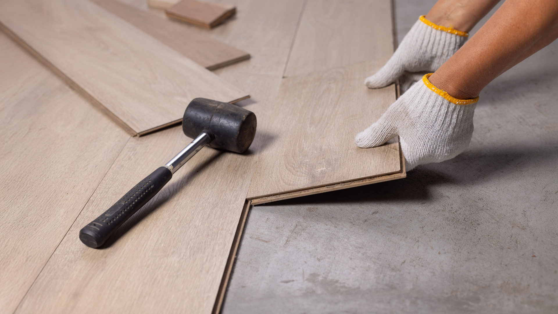 Mains dans des gants, installation d'un parquet sur une surface en béton, avec un maillet en caoutchouc à proximité.