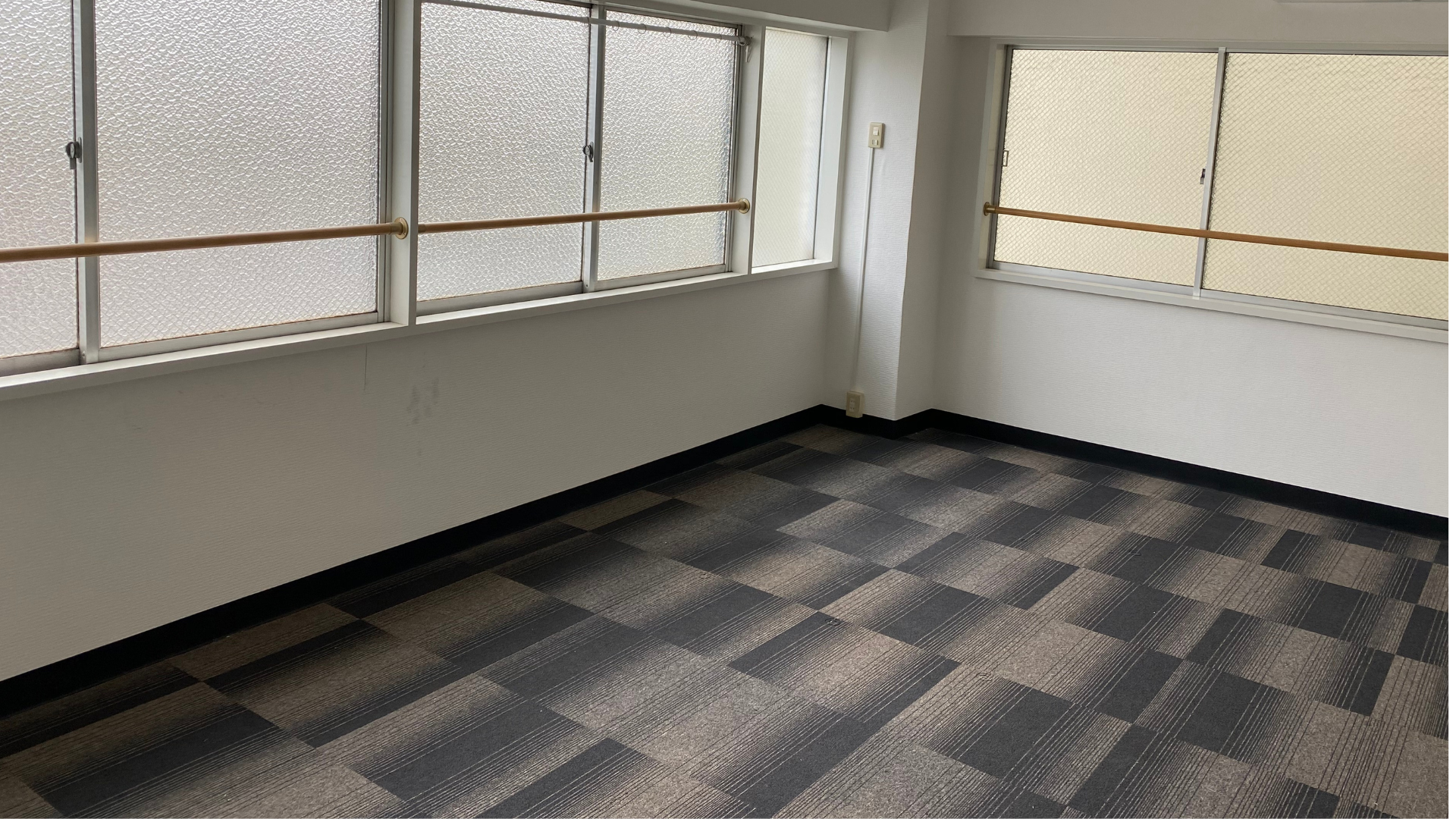 Pièce vide avec fenêtres. Moquette à carreaux gris et noir, murs blancs, boiseries marron.
