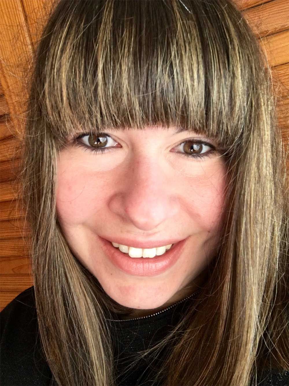 Mulher com cabelos castanhos e franja sorrindo, contra um fundo com painéis de madeira.