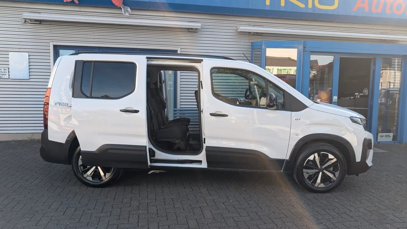 Weißer Pickup-Van mit geöffneter seitlicher Schiebetür, geparkt vor einem Autohaus.