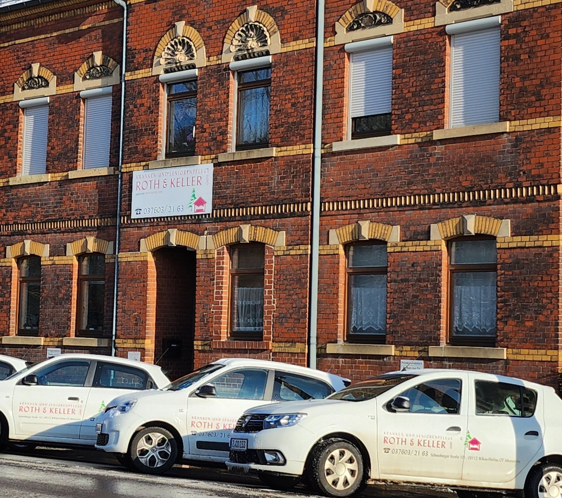 Rotes Backsteingebäude mit davor geparkten weißen Autos; auf dem Schild steht „Kuth und Sohn“.