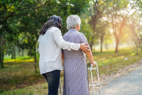 Eine Frau hilft einer älteren Person mit Gehhilfe auf einem Weg im Freien; beide blicken voneinander weg.