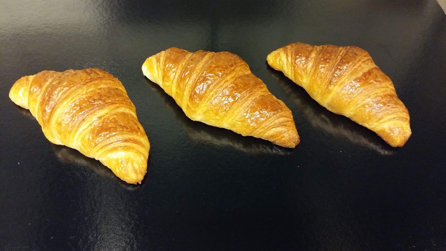Trois croissants dorés sur une surface noire, avec un léger éclat dû au glaçage au beurre de la pâte.
