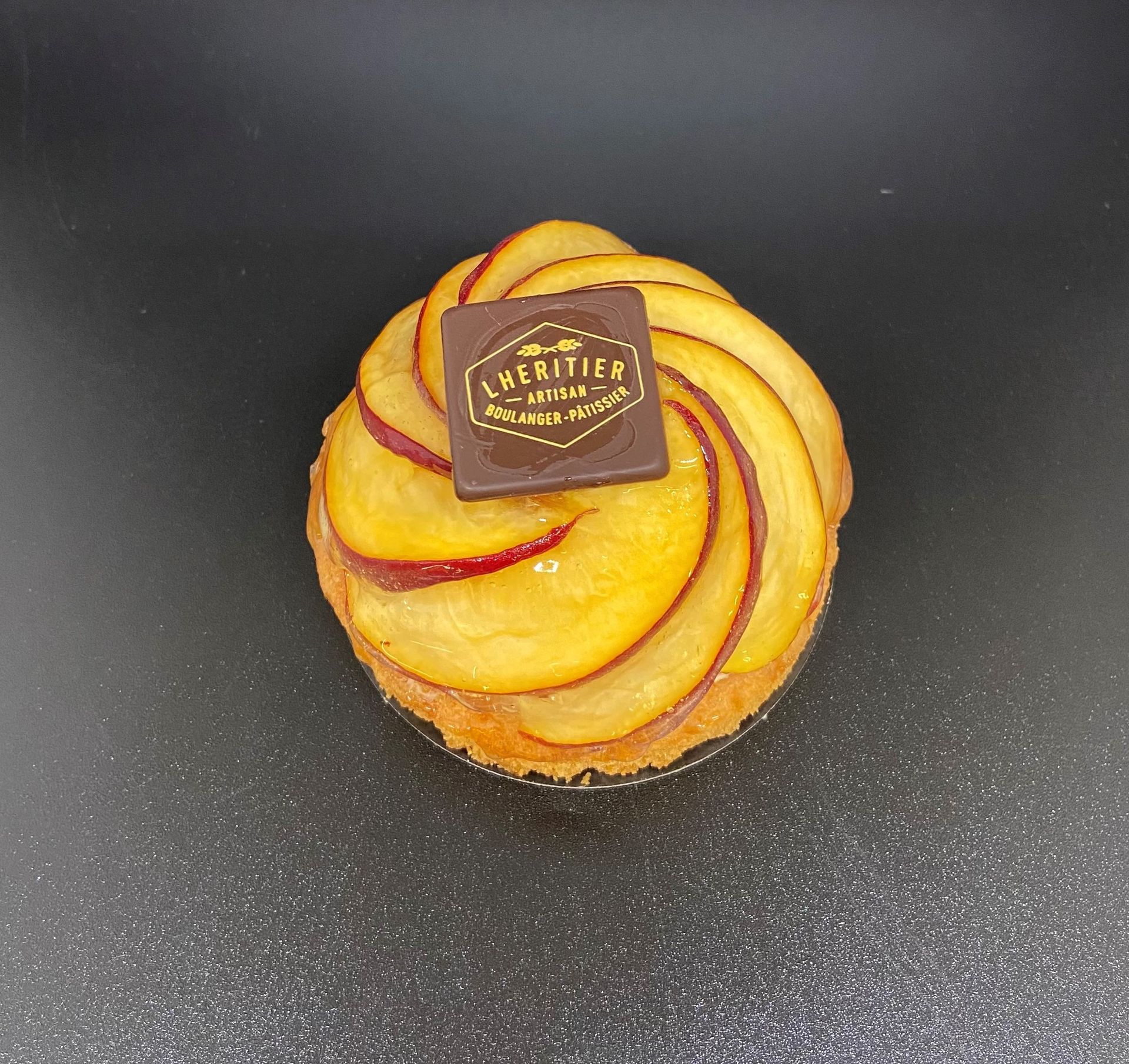 Tarte aux pêches avec des tranches de fruits disposées en spirale, surmontée d'un carré de chocolat, sur une surface foncée.