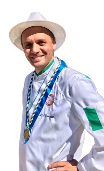 Un homme en toque et uniforme de chef sourit à l'extérieur. Il porte une médaille aux détails verts et blancs.