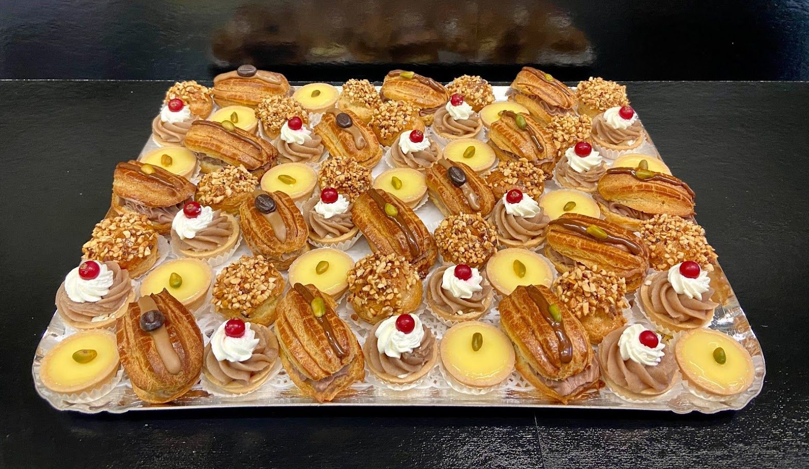 Plateau de pâtisseries colorées, glacées en rose, vert, marron et garnies de framboises et de crème.