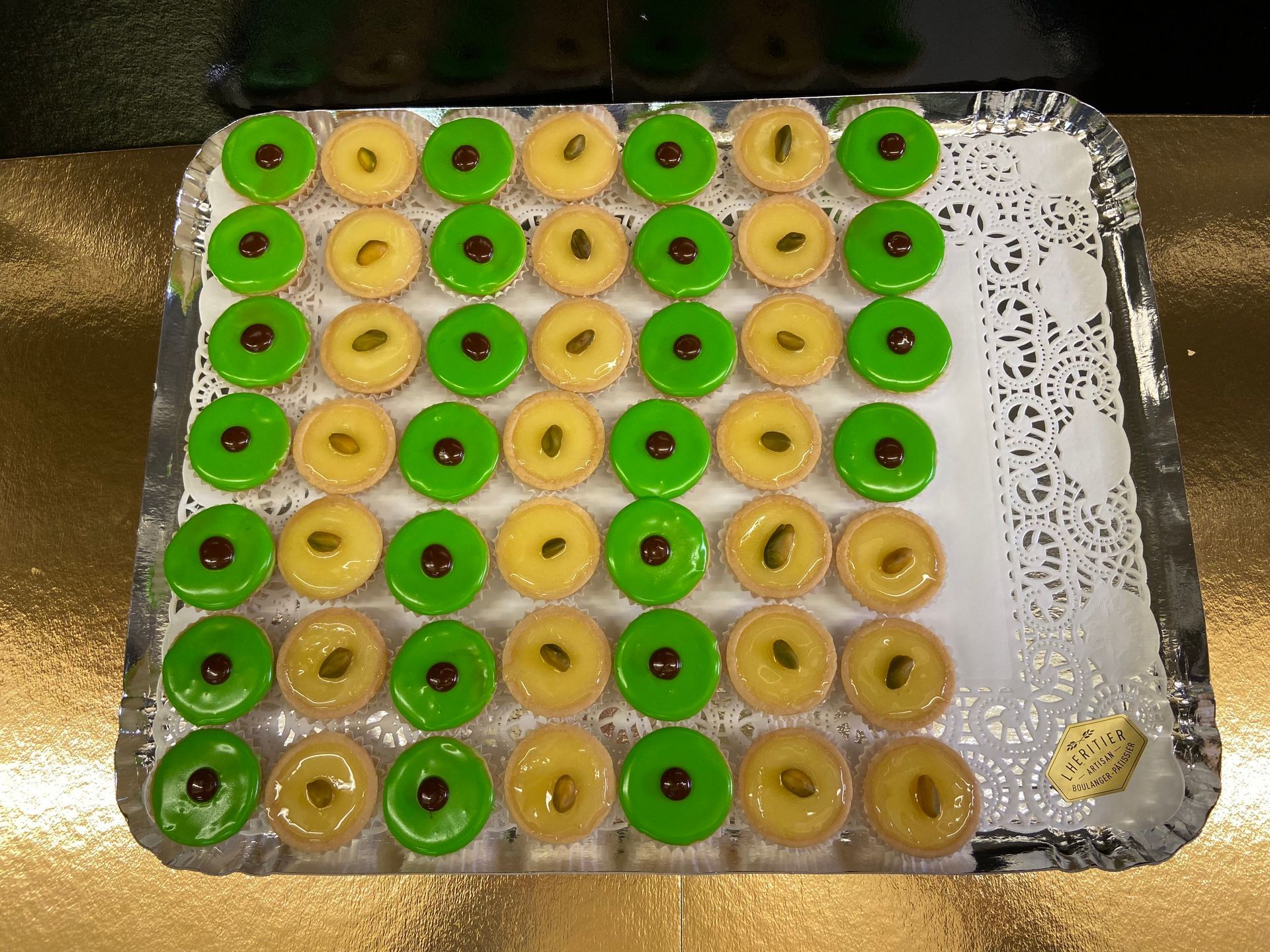 Plateau de mini tartelettes colorées : vertes et jaunes avec des points de chocolat, disposées sur une surface dorée.