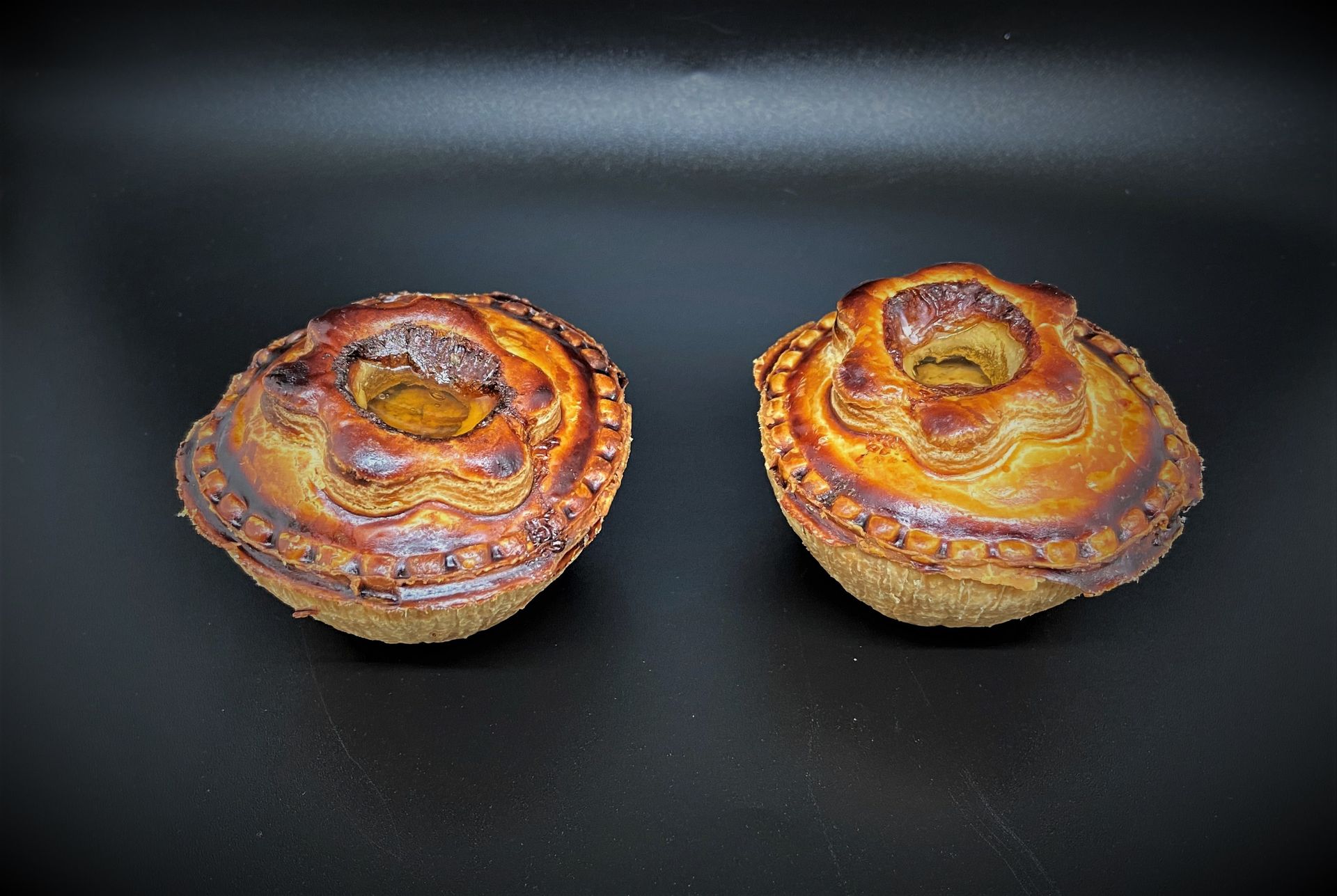 Deux tourtes à la viande rondes et dorées avec des croûtes décoratives sur un fond sombre.