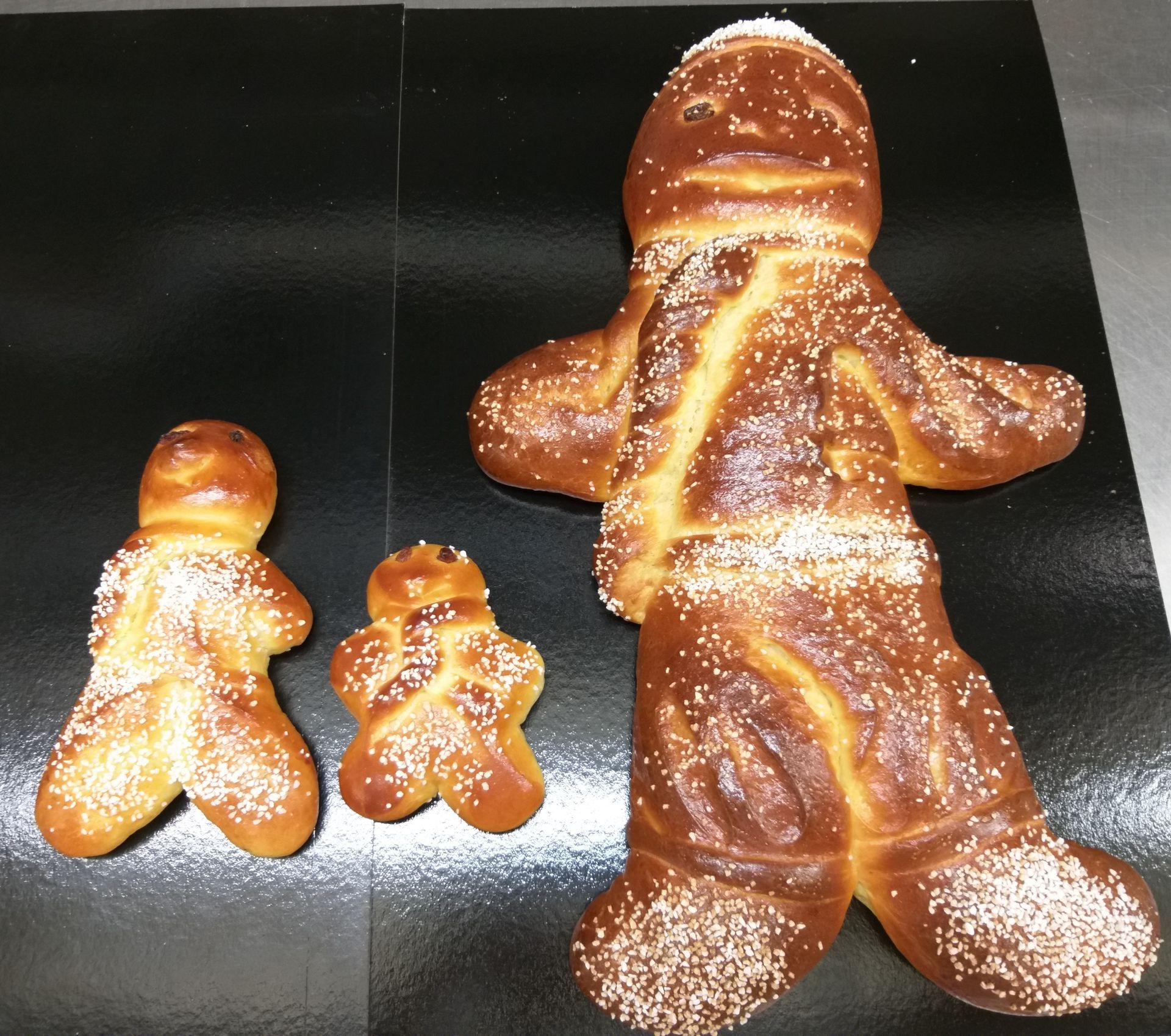 Trois figurines de pain challah de différentes tailles, en forme de personnes, sur une surface noire.