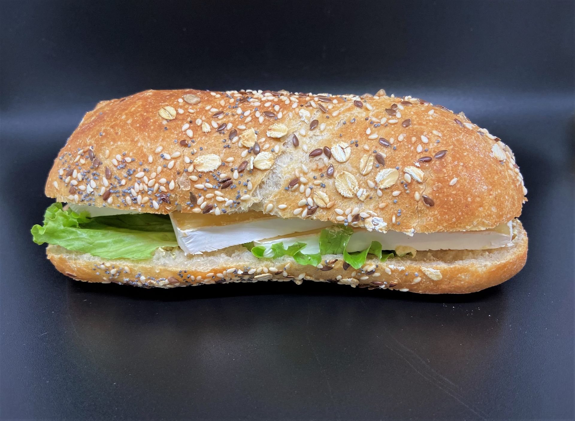 Sandwich sur un petit pain aux graines, avec de la laitue et une garniture de couleur claire, sur une surface foncée.