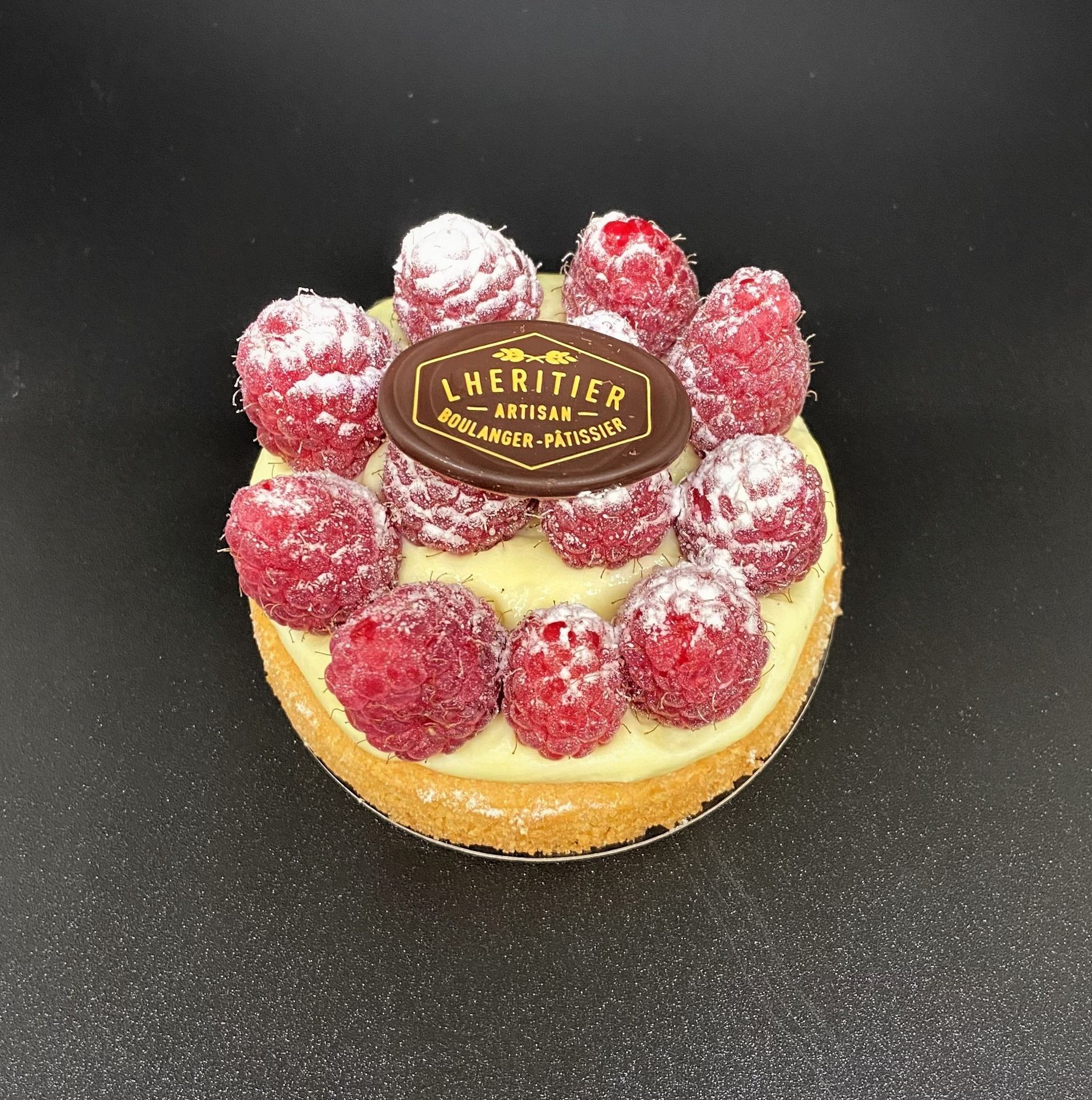 Tarte aux framboises avec sucre glace, garniture à la crème et une plaque de chocolat sur une surface noire.