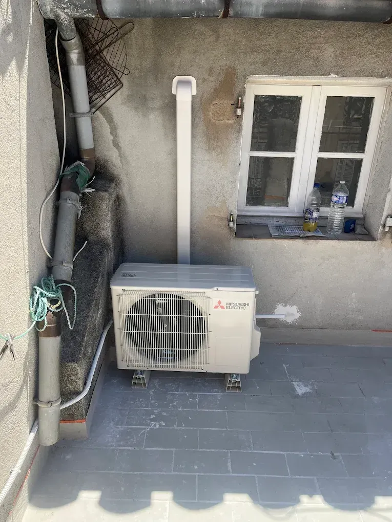 Un climatiseur Mitsubishi est installé sur une terrasse carrelée de gris, à côté d'un mur percé d'une fenêtre et de tuyauterie.