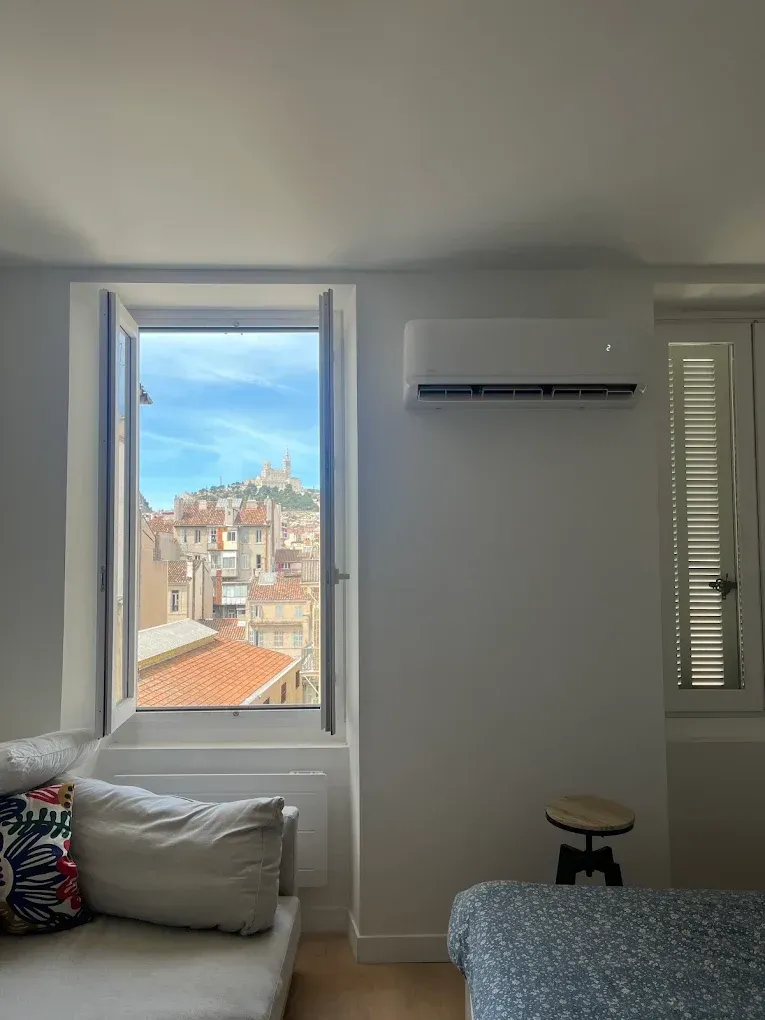 Vue depuis la fenêtre d'une chambre montrant une ville européenne avec un monument au loin, à côté d'un canapé et d'un climatiseur.