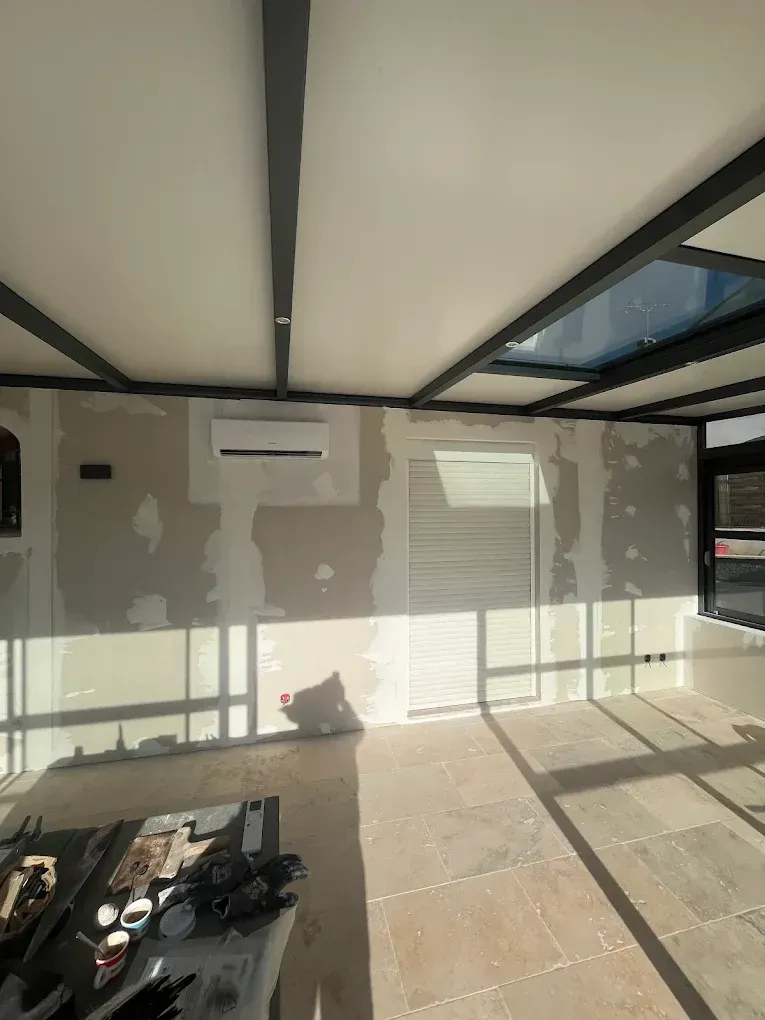 Intérieur d'une pièce en construction avec des murs en plaques de plâtre recouvertes de bandes blanches, un plafond à ossature métallique noire et un sol en carrelage clair.