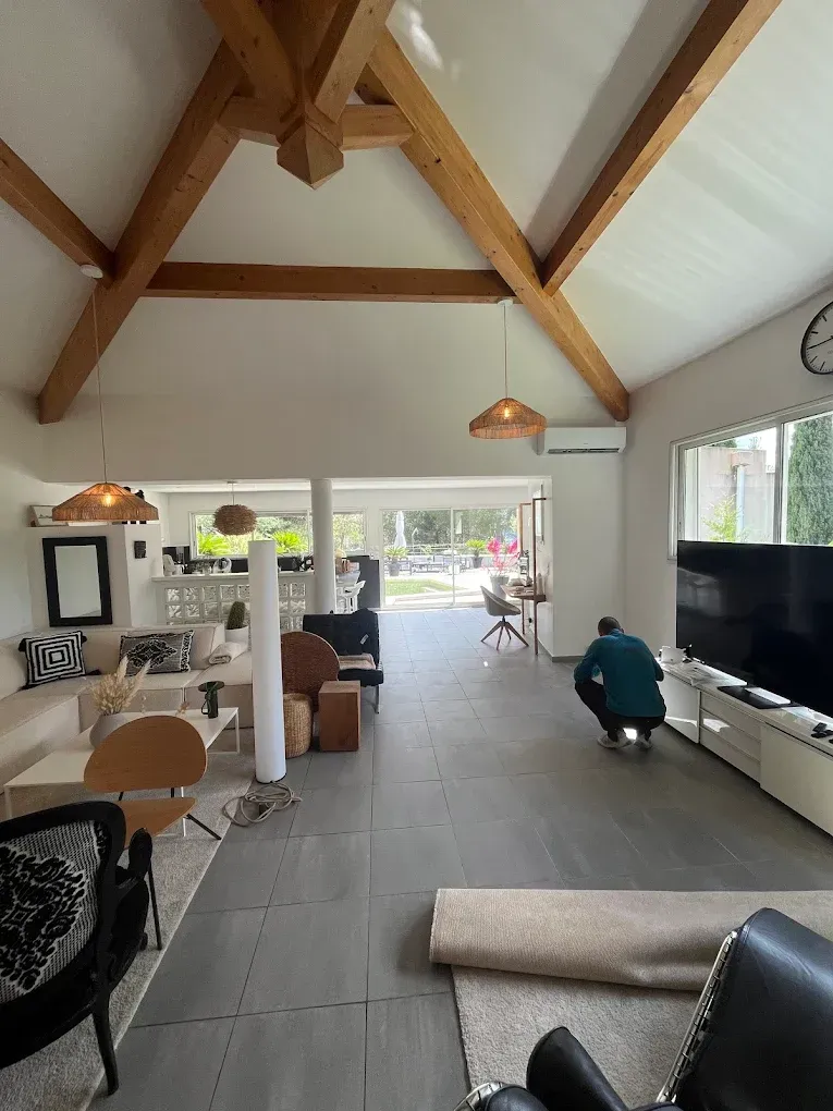 Un salon spacieux avec des plafonds voûtés à poutres apparentes, une personne accroupie près d'une grande télévision et un agencement ouvert.