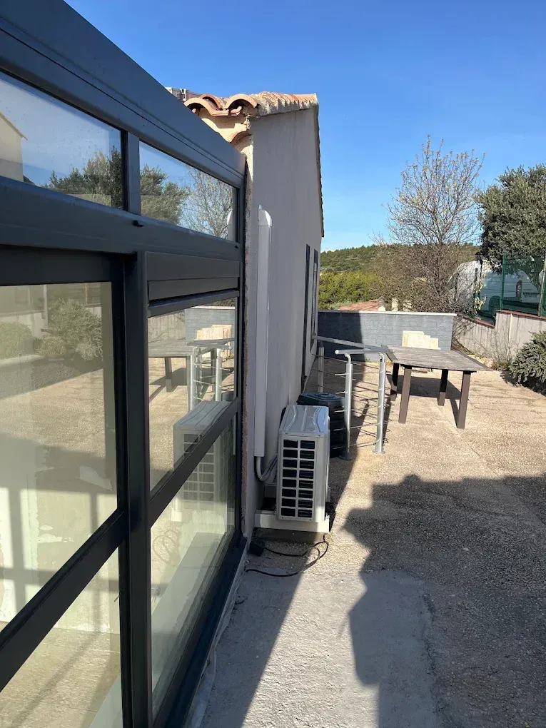 Une paroi vitrée à cadre gris s'étend le long du côté d'un bâtiment, avec un climatiseur et du mobilier de terrasse à proximité.