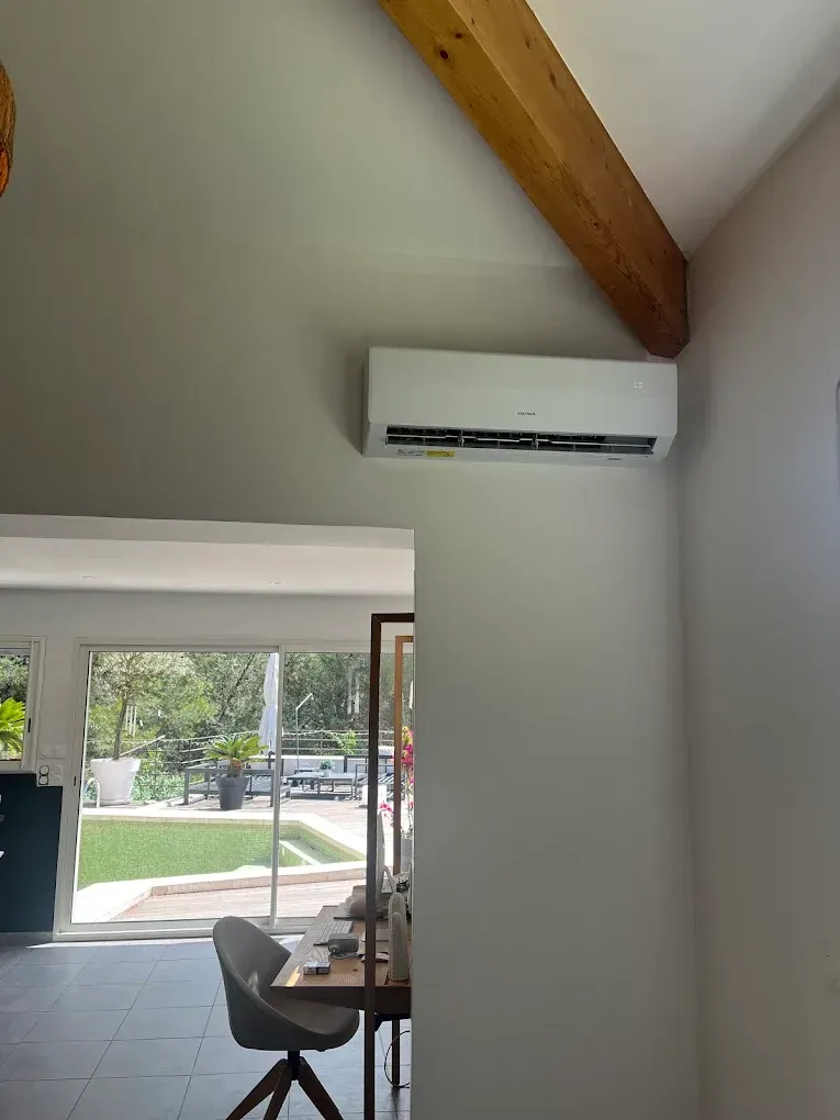 Un climatiseur mural blanc installé dans une pièce avec une poutre apparente au plafond et une vue sur un patio extérieur.