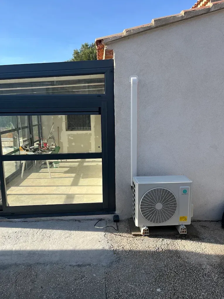 Une unité de pompe à chaleur extérieure fixée contre un mur extérieur clair, à côté d'une véranda moderne aux parois de verre à cadre foncé.