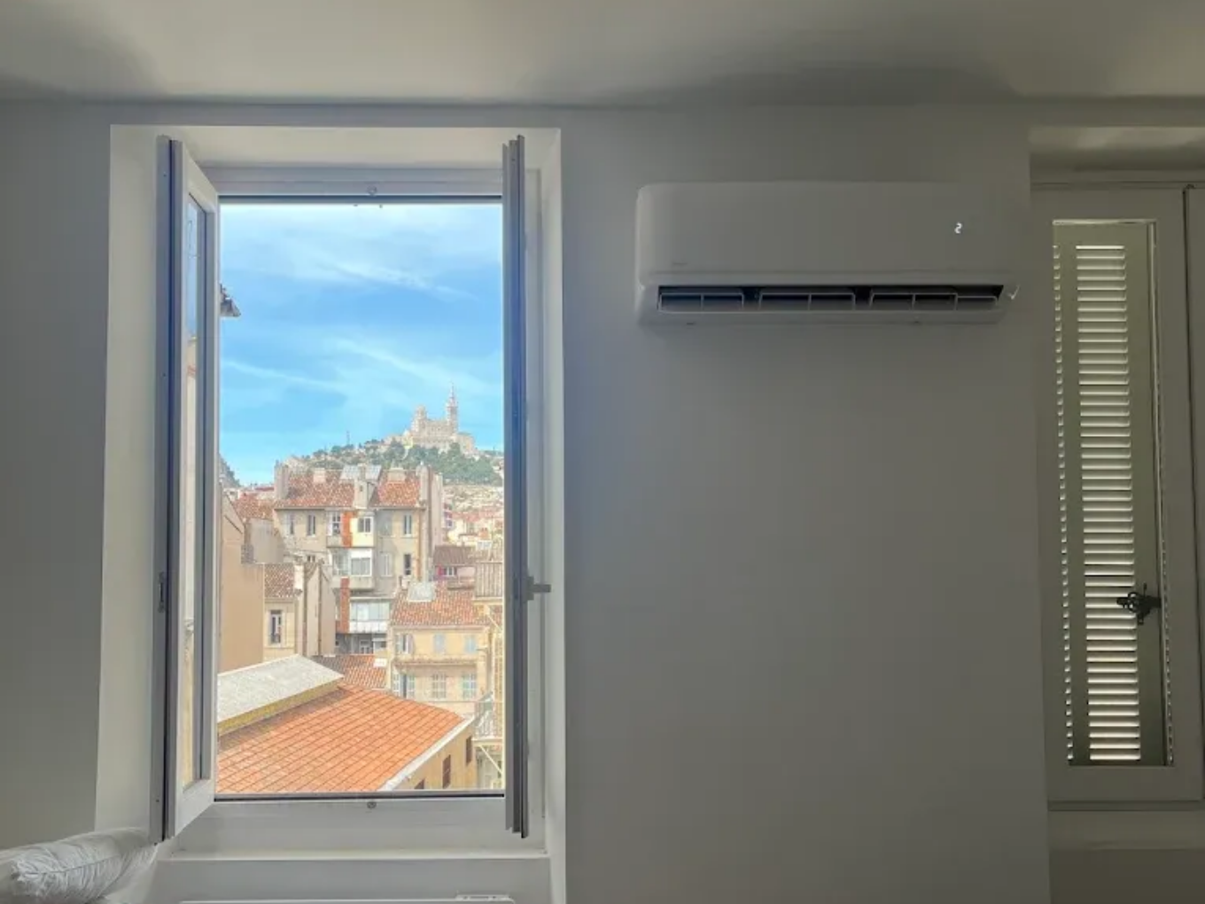 Une fenêtre ouverte donne sur une ville européenne avec une basilique perchée sur une colline, à côté d'un climatiseur mural blanc.