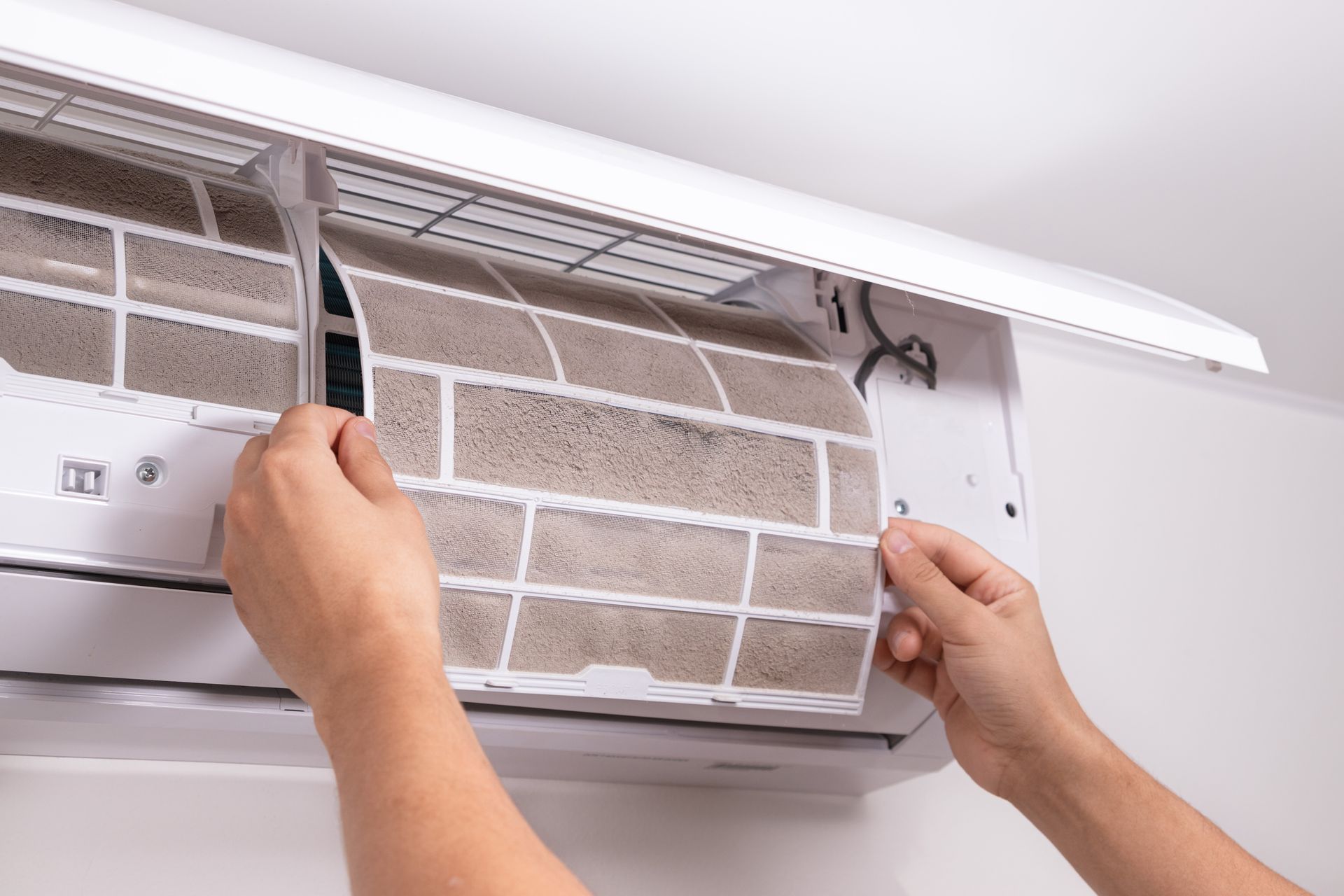Deux mains retirent un filtre à mailles poussiéreux d'un climatiseur mural blanc pour le nettoyer.