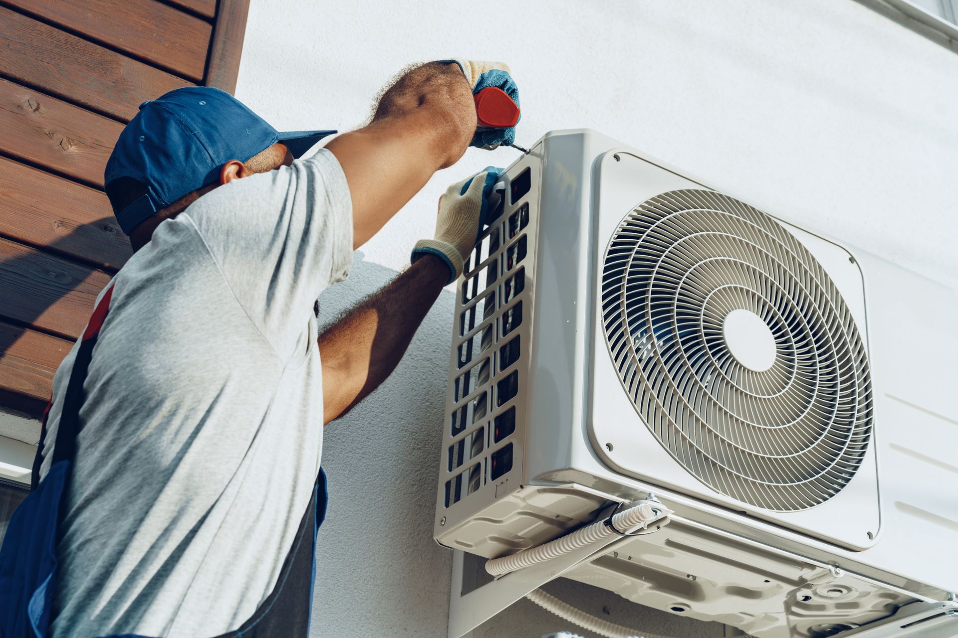Un technicien, coiffé d'une casquette bleue et vêtu d'une chemise de travail, répare une unité de climatisation extérieure fixée au mur d'un bâtiment.