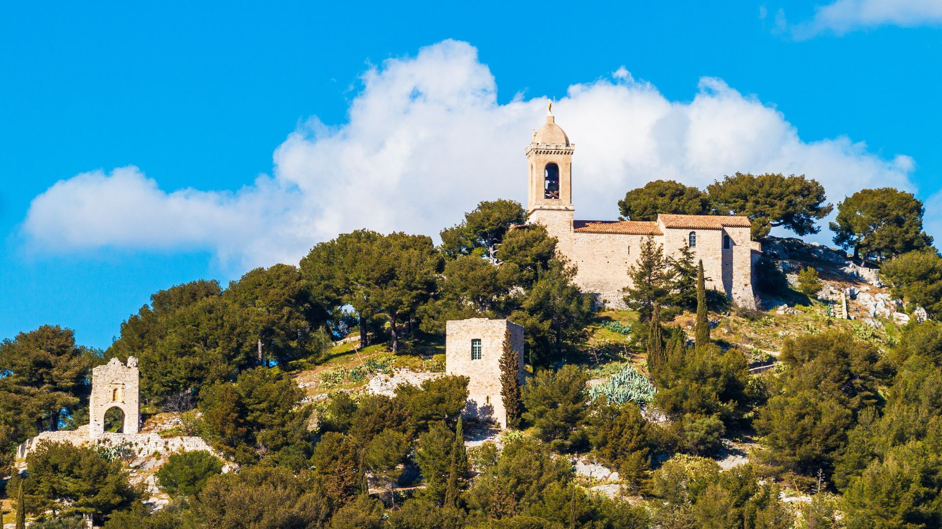 Une église en pierre, avec son clocher et des ruines à proximité, se dresse au sommet d'une colline boisée, sous un ciel bleu parsemé de nuages ​​blancs.