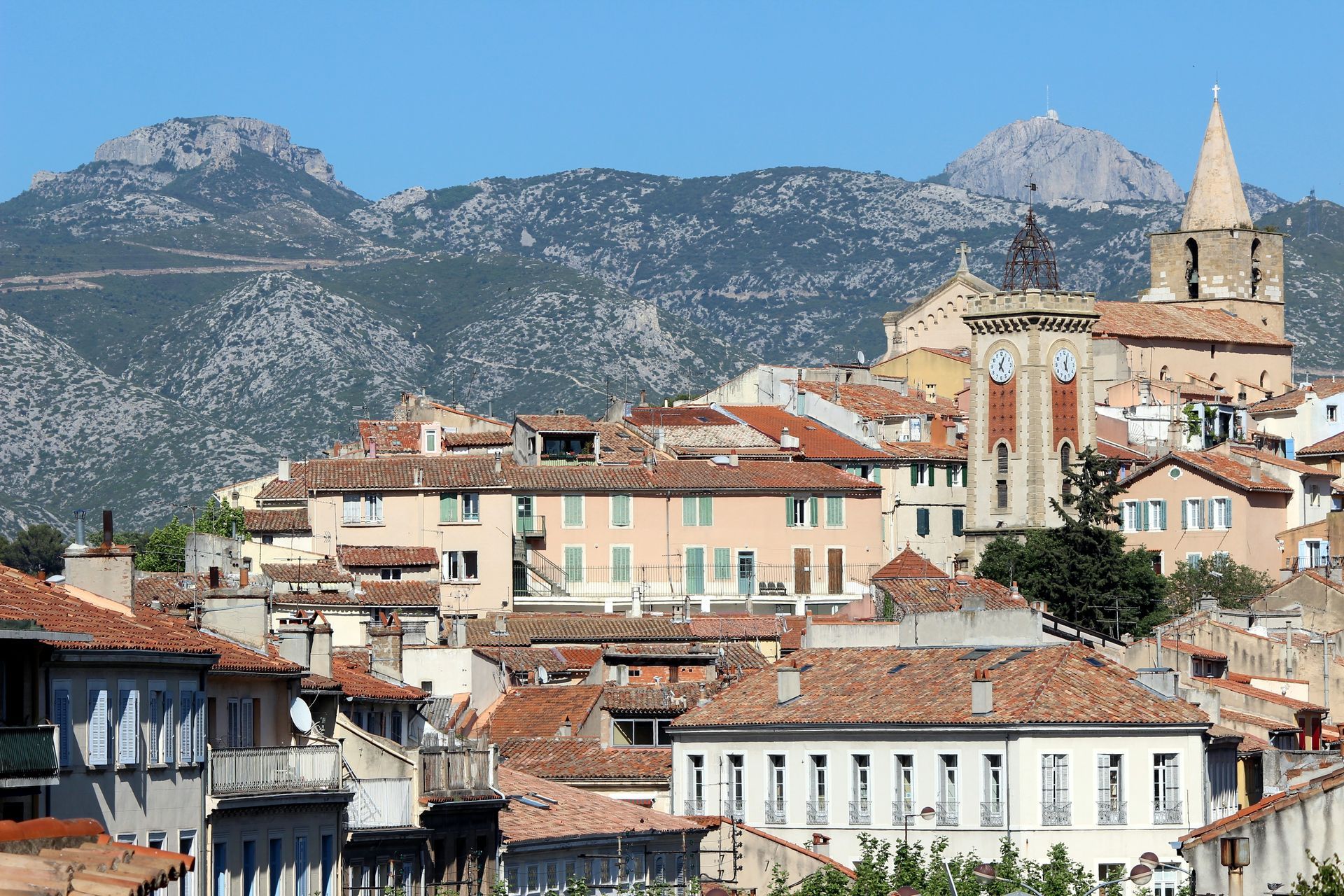 Une ville historique aux toits de tuiles rouges et à l'imposante tour de l'horloge, se détachant sur un fond de montagnes escarpées et boisées.