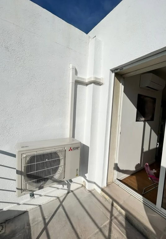 Un groupe de climatisation extérieur Mitsubishi blanc est installé sur un balcon près d'une porte ouverte.