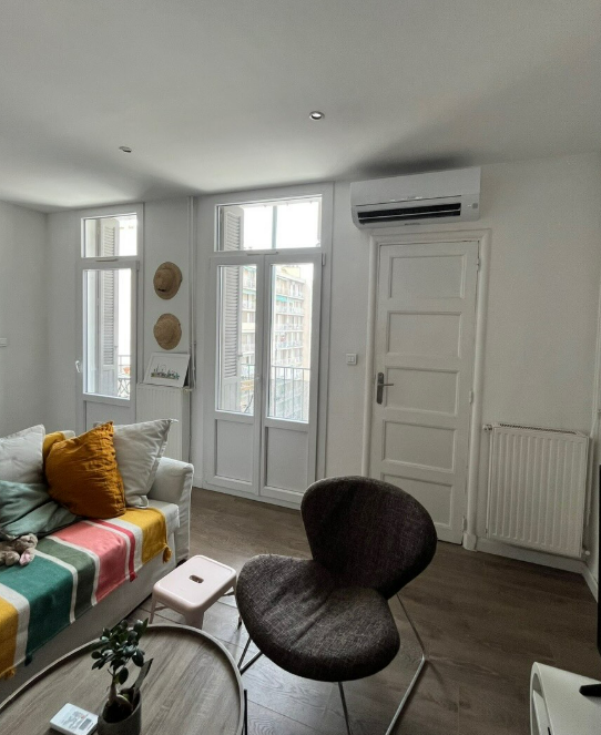 Un salon lumineux avec un canapé recouvert d'un plaid à rayures, un fauteuil foncé, une table basse et des portes blanches donnant sur un balcon.