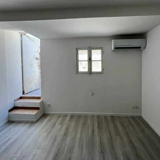 Une pièce avec un sol clair imitation bois, des murs blancs, une petite fenêtre centrale, la climatisation et quelques marches menant à une porte.