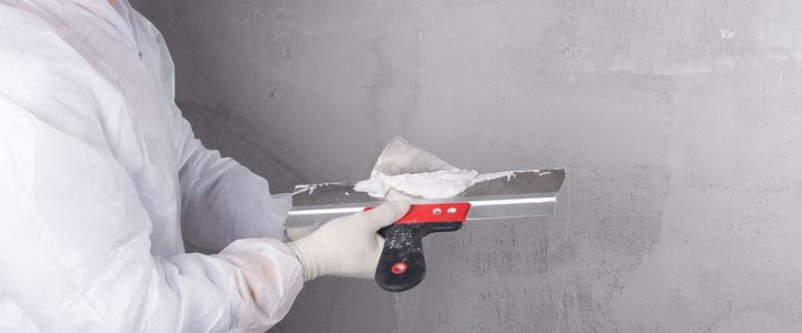 Eine Person in Schutzkleidung trägt mit einem Spachtel Gips an einer Wand auf.