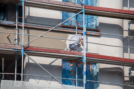 Ein Bauarbeiter auf einem Gerüst verputzt ein Gebäude. Die Fenster sind mit blauer Folie verklebt.